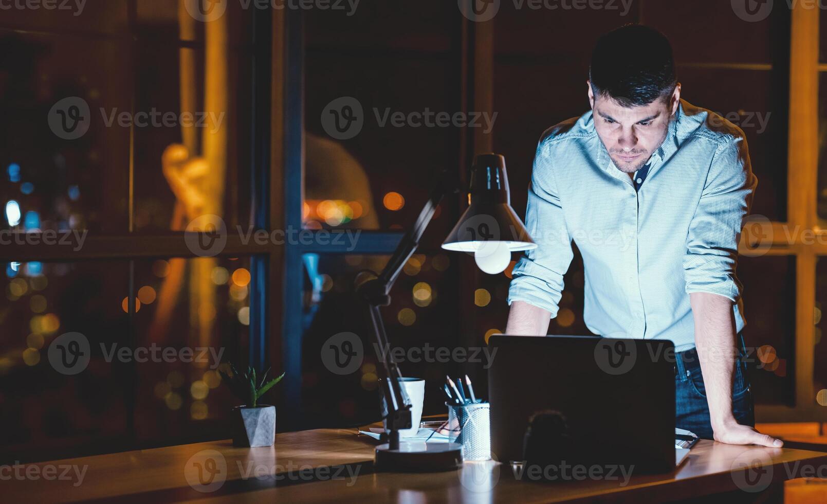 Young Entrepreneur Guy Standing Working On Laptop In Modern Office At Night. Empty Space, Low Light photo