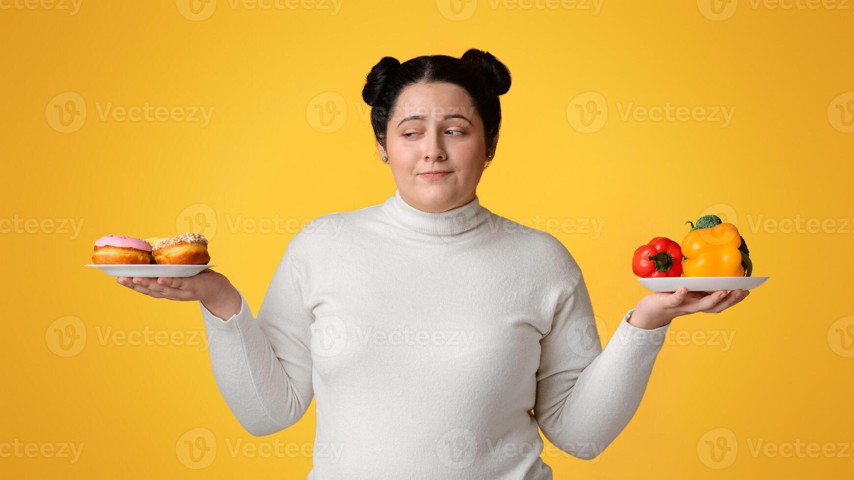 Healthy Vs Unhealthy Food. Young obese girl choosing between fresh vegetables and donuts, keeping hands as a scales over yellow background, copy space photo