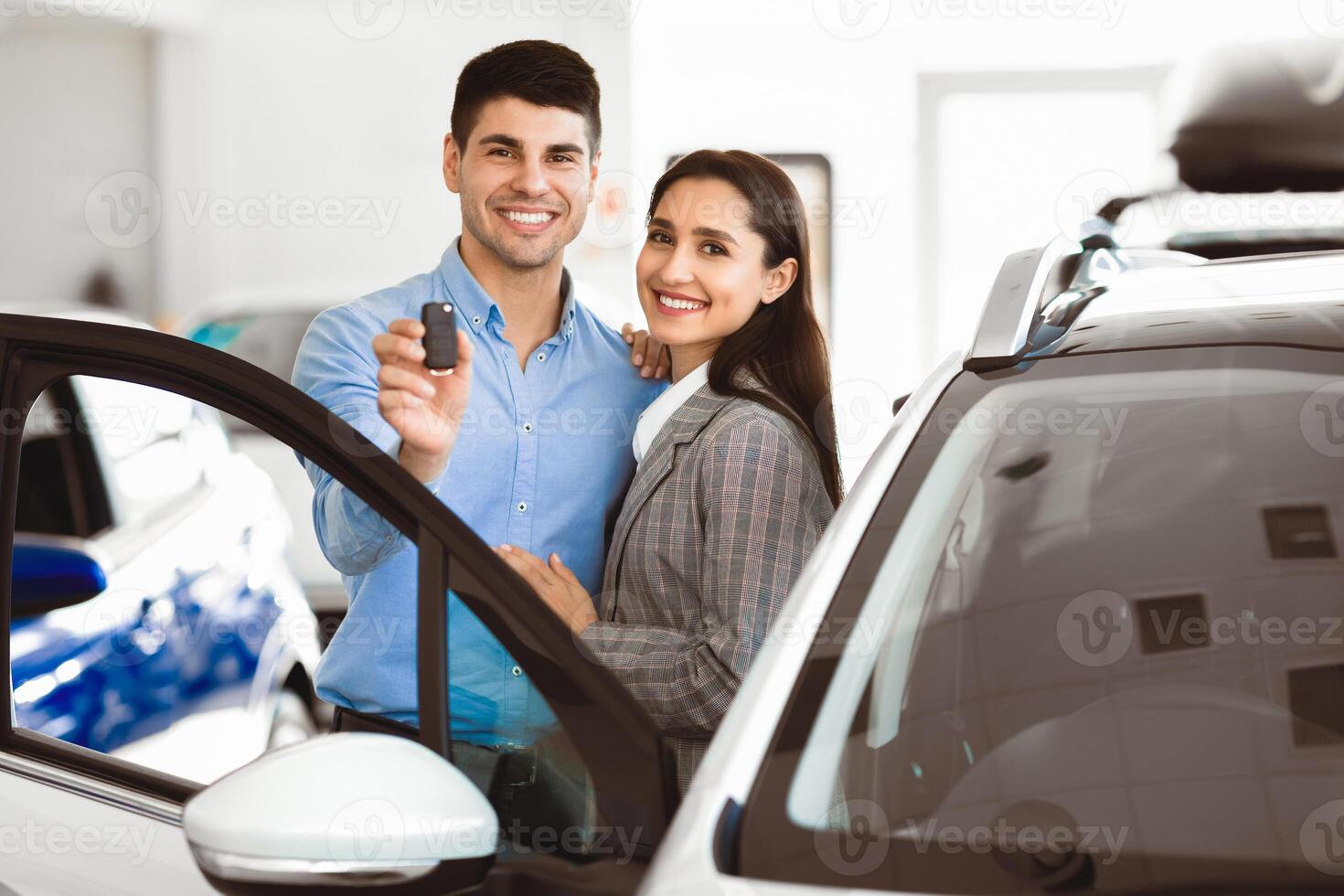 First Family Car Concept. Lovely couple hugging after buying auto together in dealership, man showing key on camera photo