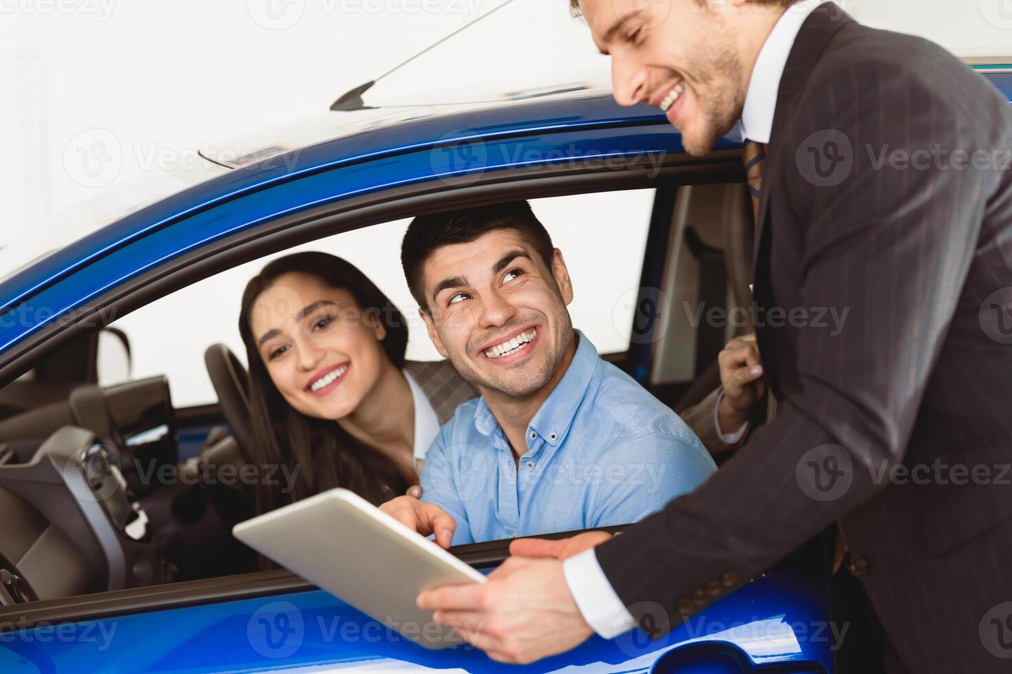 Check Out The Characteristics. Happy couple sitting in salon of new car, vendor showing tablet to them photo
