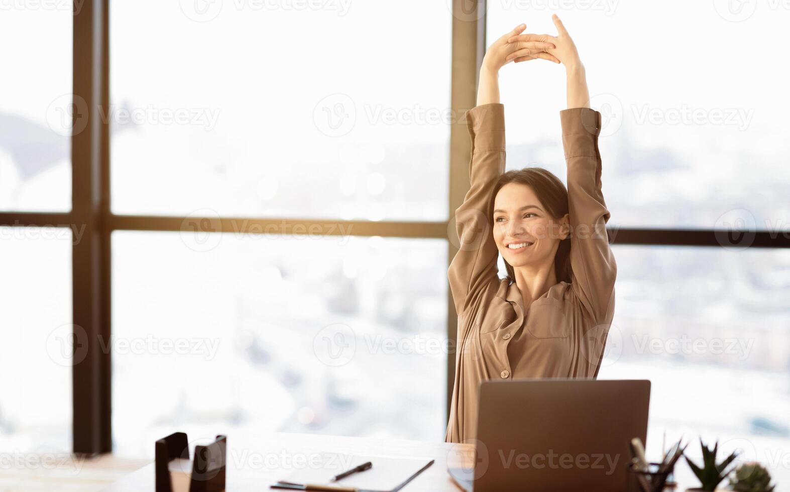 Smiling Young Woman In Brown Long Sleeve Shirt Stretching In Front Of Desk At Office, Looking Aside, Free Space photo