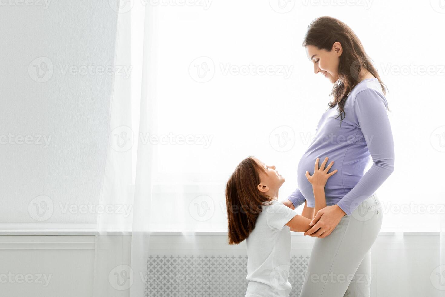 Excited little daughter touching pregnant mom tummy, standing next to window, copy space photo