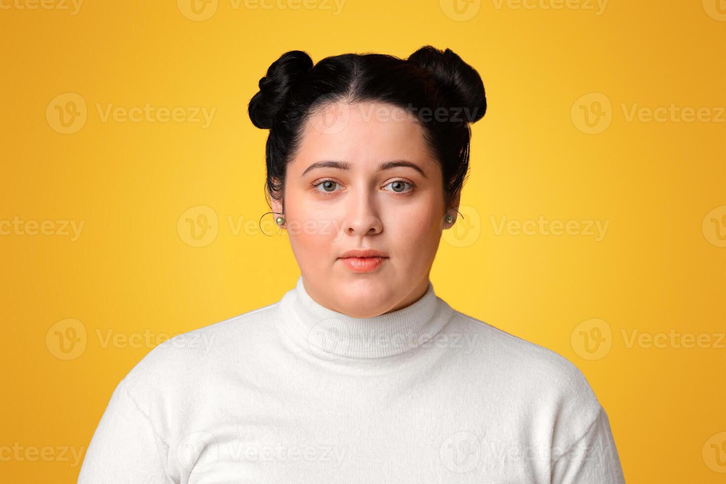 Portrait of young plus size girl with double bun hairstyle standing over yellow background and looking at camera, free space photo
