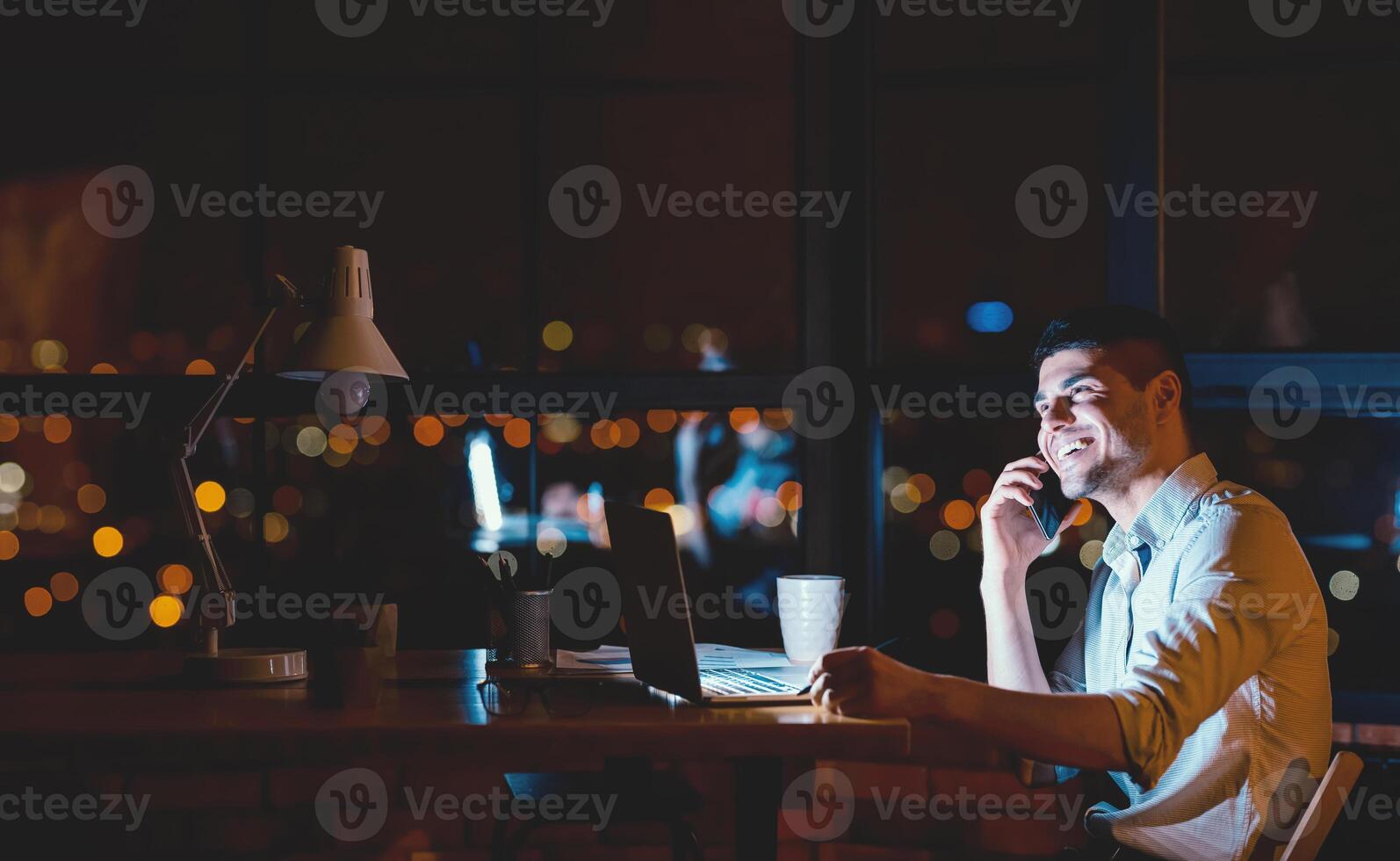 Business Calls. Cheerful Man Talking On Phone Working On Laptop Computer Sitting In Modern Office At Night. Empty Space photo