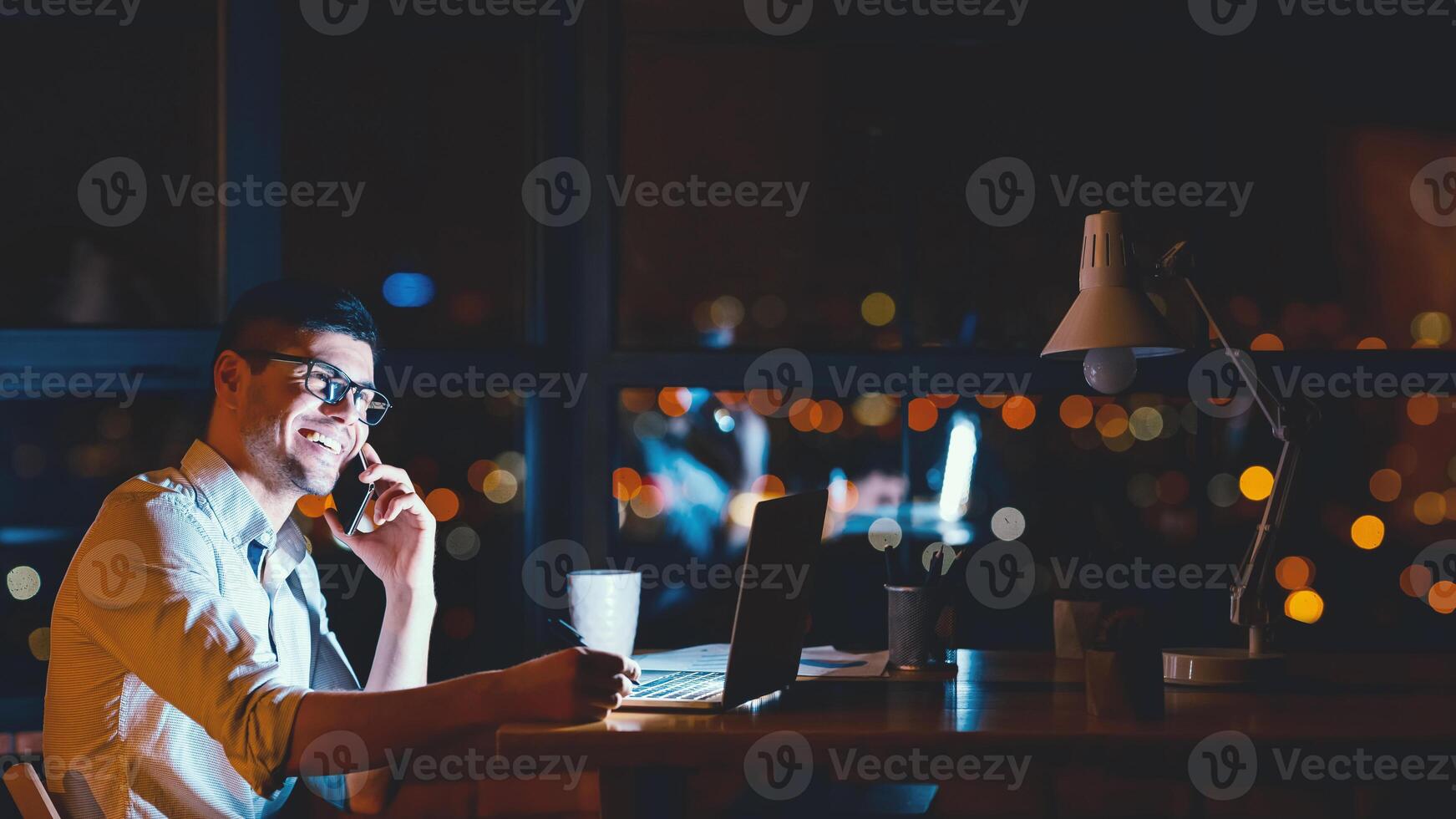 Happy Entrepreneur Guy Talking On Cellphone Sitting At Laptop In Modern Office At Night. Business People Lifestyle. Panorama, Free Space photo