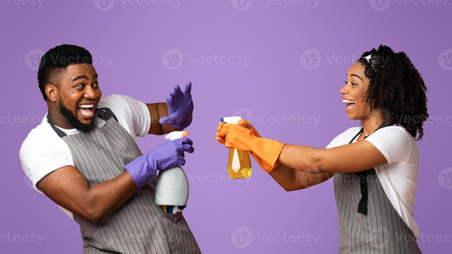 Cheerful Afro Couple In Aprons Playing With Spray Detergents, Playfully Aiming At Each Other While Posing On Pink Background In Studio photo