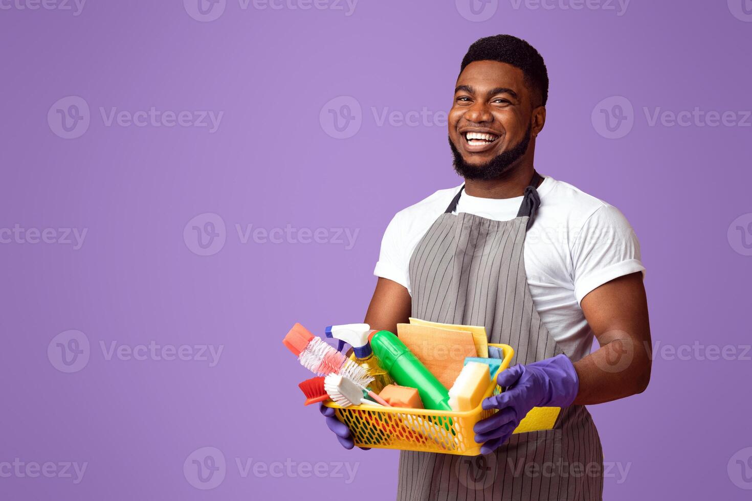 Professional Cleaner. Positive black cleaning service worker holding basket with household supplies and smiling at camera, pink background photo