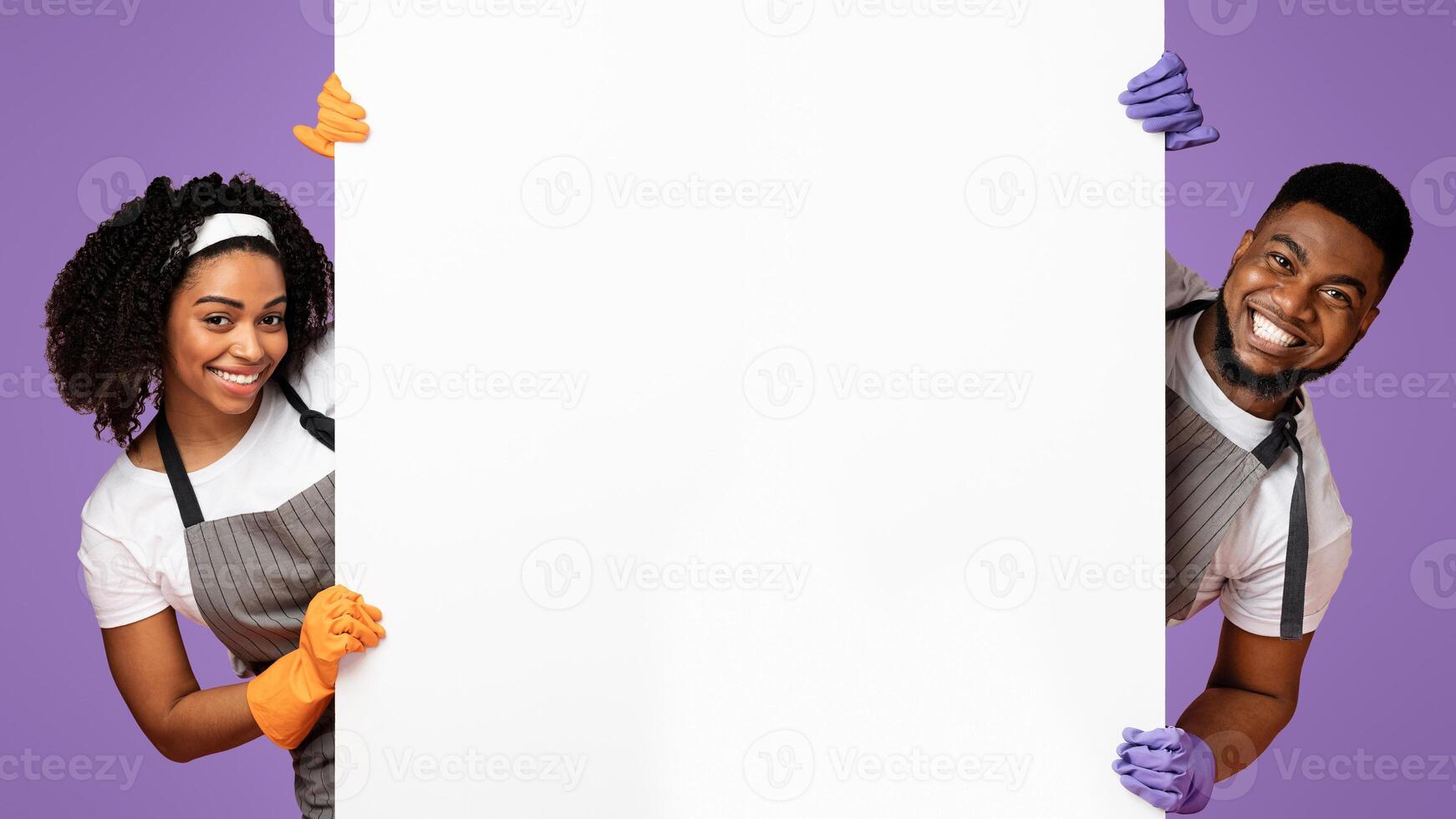 Cleaning Services Ad. Smiling black couple in gloves and aprons peeking out of behind blank white advertising board, panorama with copy space photo