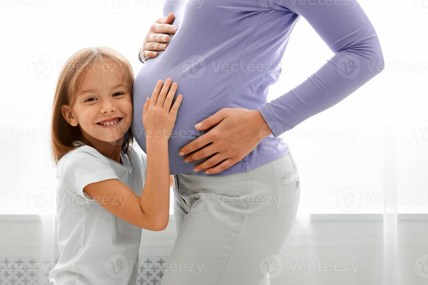 Close up of happy little daughter embracing her pregnant mom, home interior photo