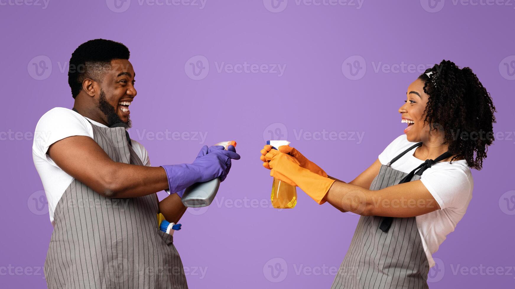 Household Fun. Joyful black couple fooling during spring-cleaning, aiming detergent sprayers at each other, standing over pink studio background photo