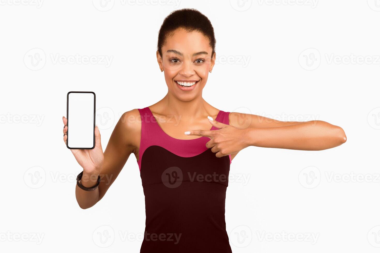 Great Fitness App. Happy Afro Woman Showing Smartphone Empty Screen Pointing Finger Standing Over White Studio Background. Mockup photo