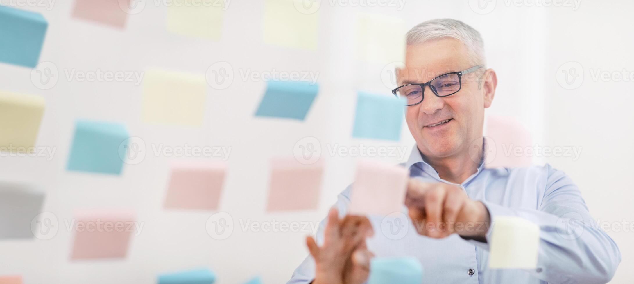 Paper Reminder. Smiling senior male manager checking sticky notes glued to glass wall, free space photo