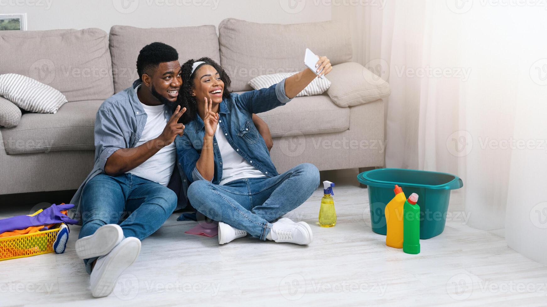 Happy Black Couple Taking Selfie On Smartphone After Tidying Flat Together, Relaxing On Floor After Spring-Cleaning, Showing Peace Gestures photo