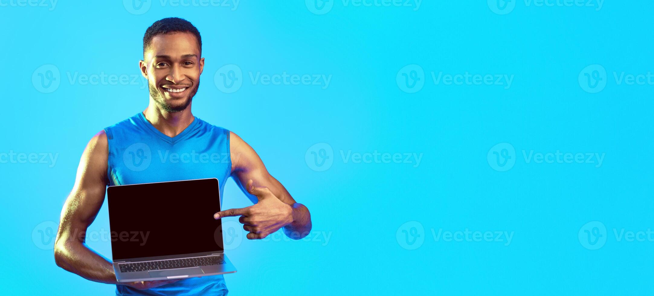 Online Gym Concept. Fit African Man Showing Laptop With Empty Screen For Your Text On Blue Studio Background. Mockup, Panorama photo