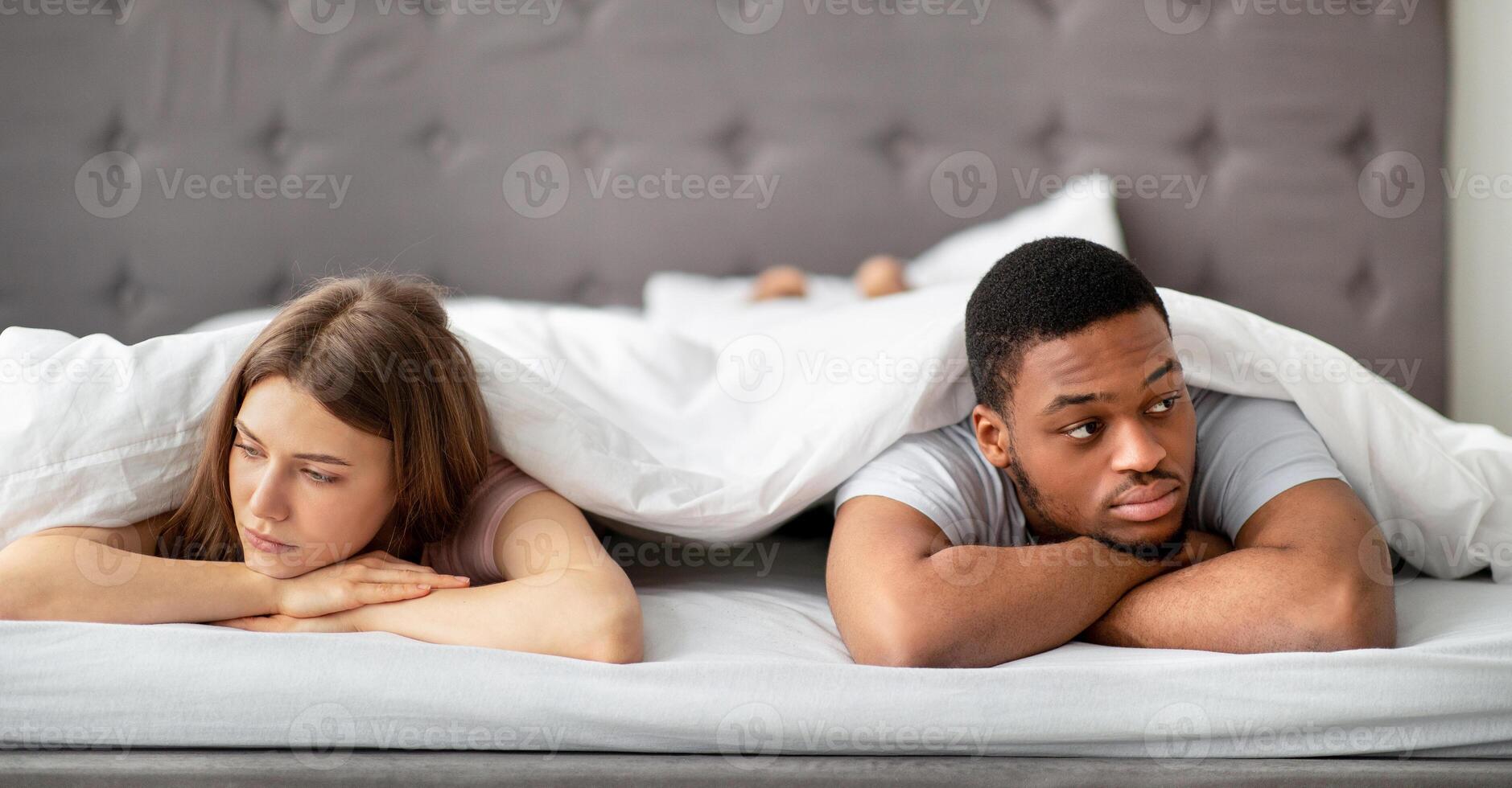 Sad Caucasian woman and her black husband lying apart on bed at home, looking in opposite directions. Interracial couple feeling offended after fight, suffering from relationship or sexual problems photo