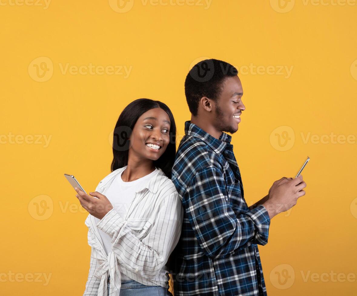 Blogs, social media, unusual message and interesting event. Cheerful young african american lady look over shoulder in smartphone of smiling husband, isolated on yellow background, studio shot photo