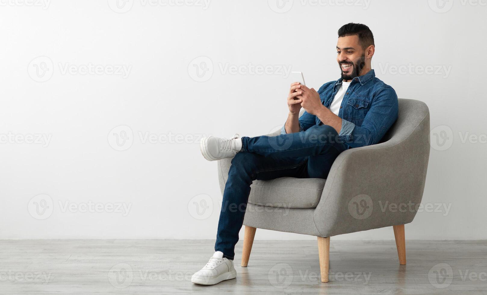 Joyful young Arab guy sitting in armchair with smartphone, working remotely or chatting against white studio wall, empty space. Middle Eastern man watching movie, having online meeting photo
