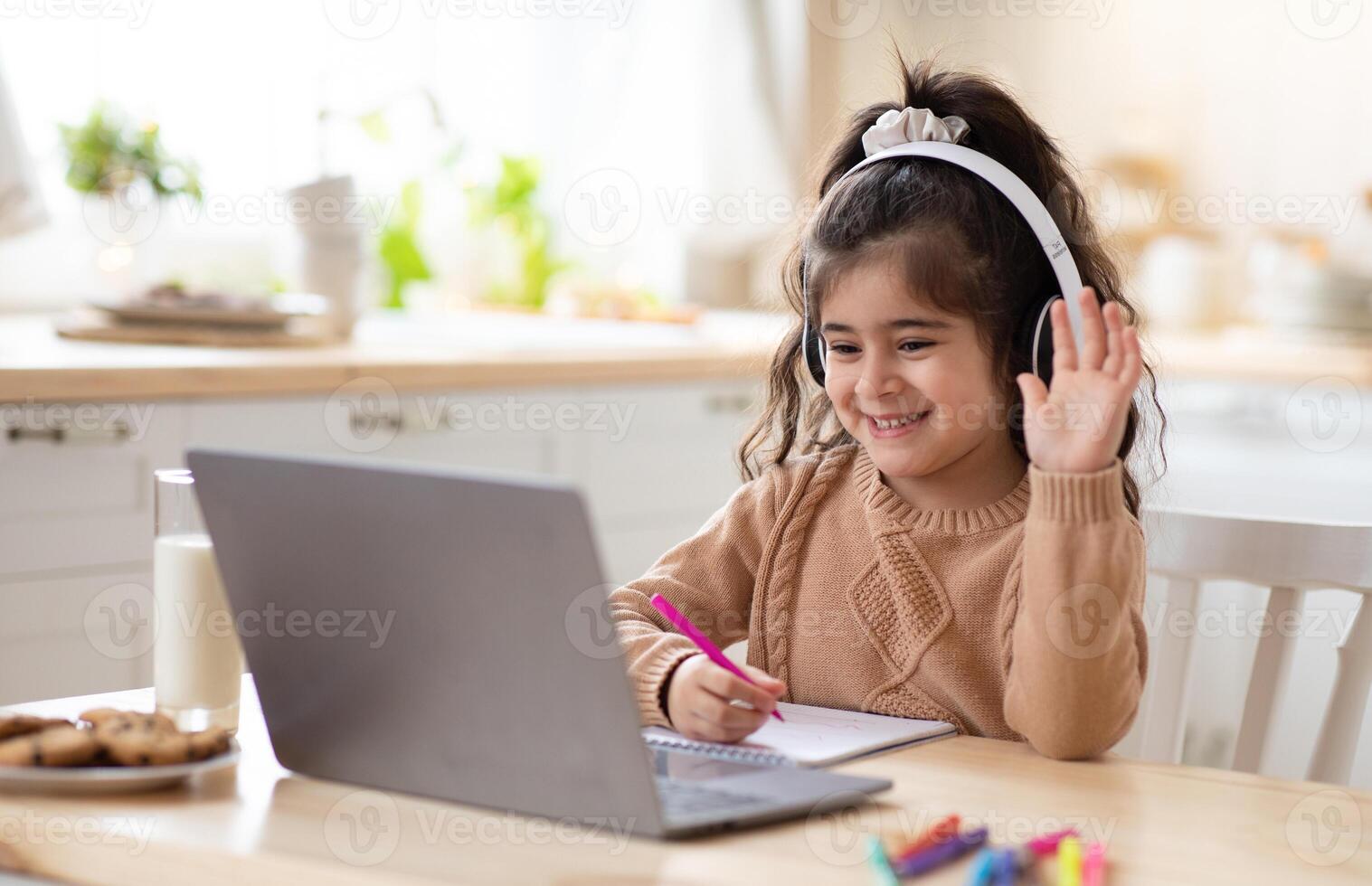 Distance Learning. Cute little home schooler girl having lesson with laptop in kitchen, studying with computer during coronavirus quarantine, enjoying online education, waving hand at camera photo