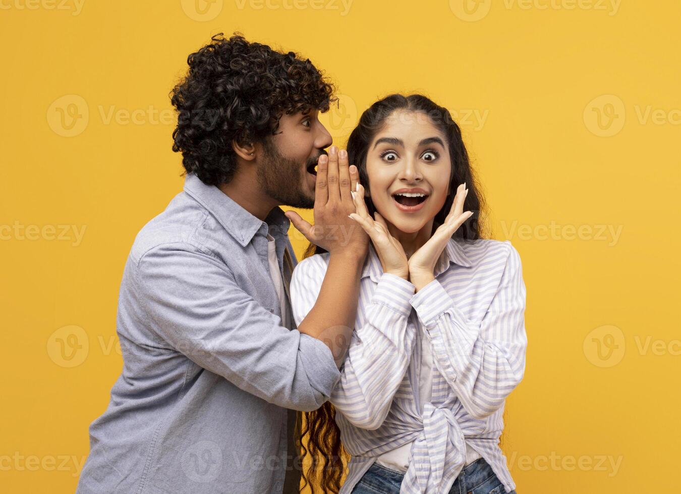 Shock, gossip, share advice. Young indian man whispering to lady on ear, happy woman with open mouth excited and surprised, posing on yellow studio background photo