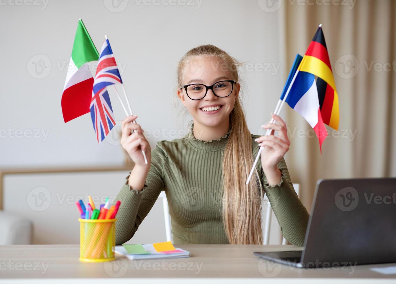 Cheerful blonde teen girl with glasses sitting at workdesk with international flags, home interior, using laptop, studying foreign languages. International education, online language courses photo