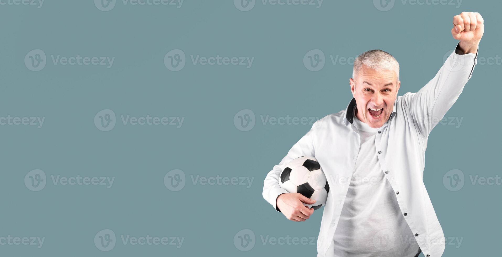 Celebrating Goal. Cheering senior man posing with soccer ball, making winner gesture, blue studio wall photo
