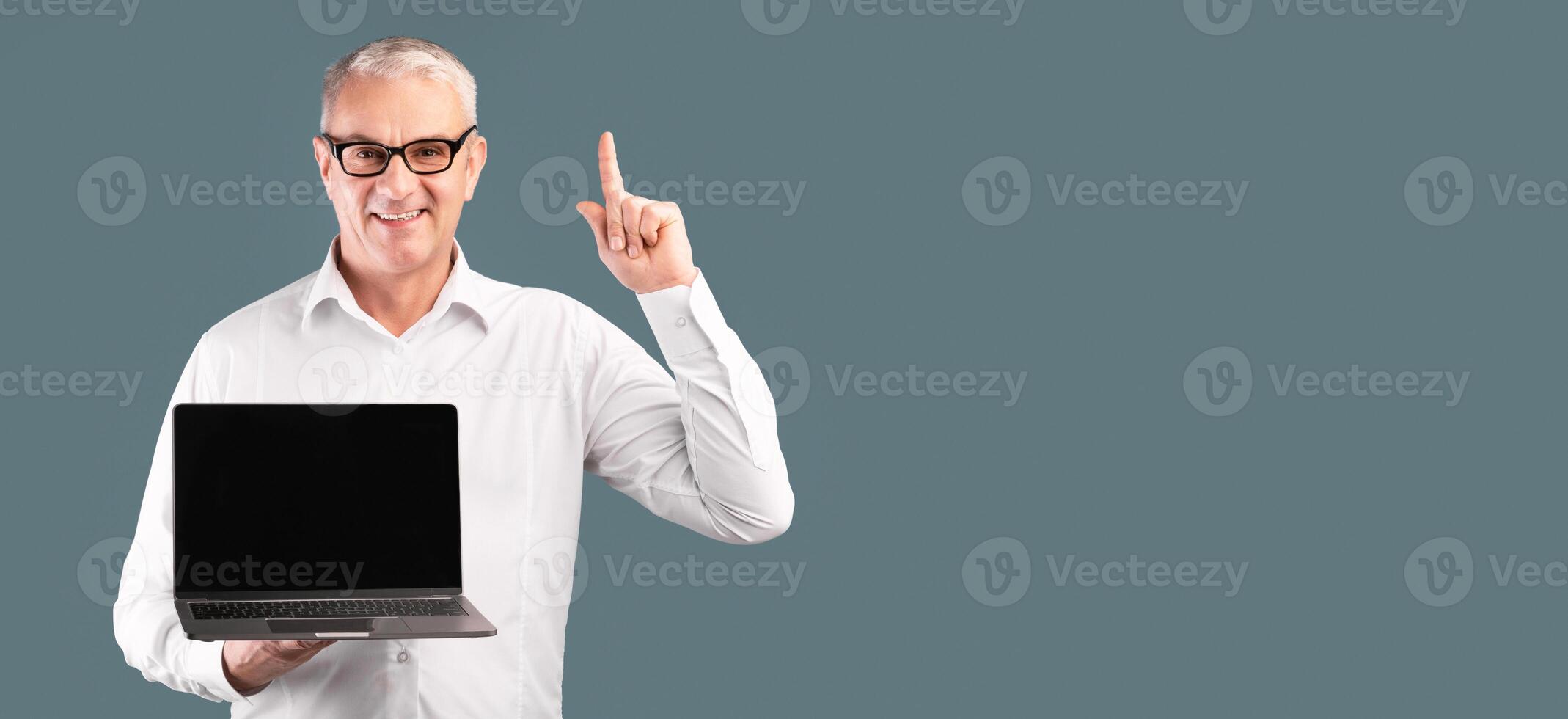 Useful Website. Middle-aged man pointing up and holding laptop with blank black screen over blue studio wall, mock up photo