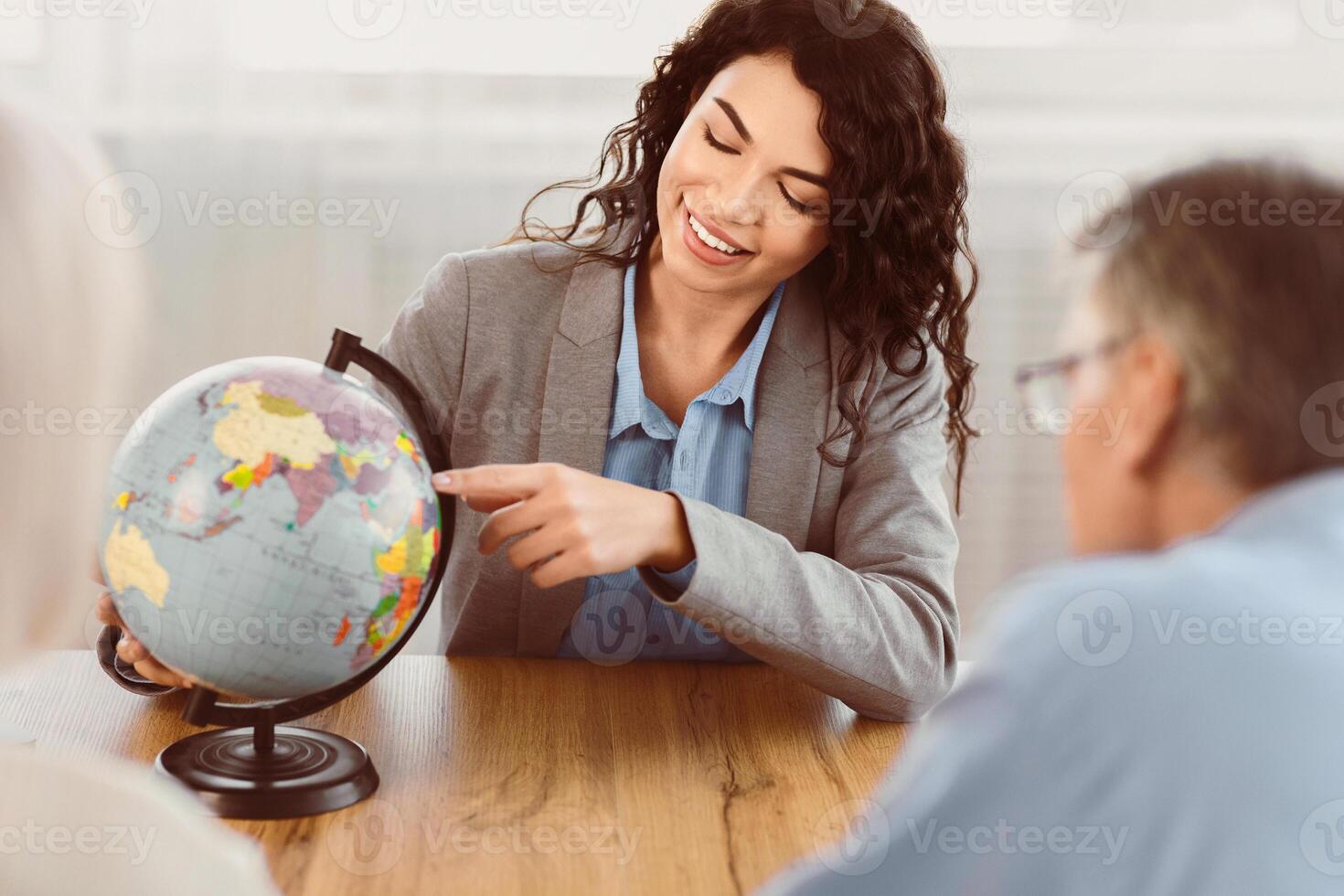 Choosing Trip. Beautiful mexican agent proposing last minute travel offer to mature couple, pointing at globe, copyspace photo