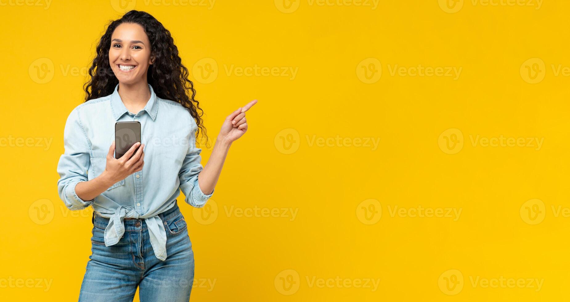 Special Offer. Portrait of excited young lady pointing finger aside at free copy space holding using cell phone isolated on yellow orange wall. Positive female showing empty mockup place for banner ad photo