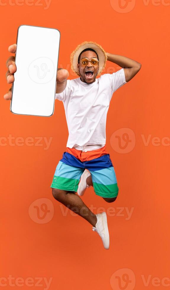 Cool mobile offer. Excited jumping black guy demonstrating smartphone with white screen on orange studio background, collage with mockup copy space for app or website design photo