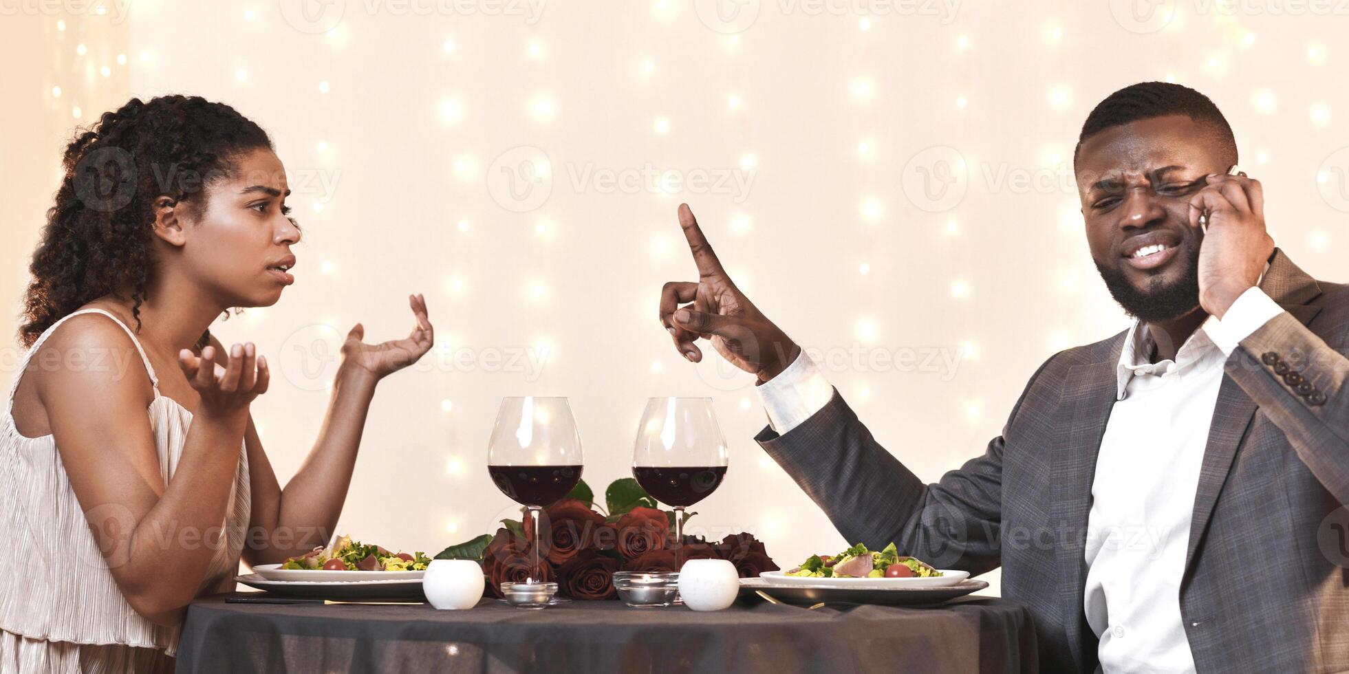 Annoyed afro woman looking at her man talking by phone while having date in restaurant, conflicts in couple concept, panorama, side view photo
