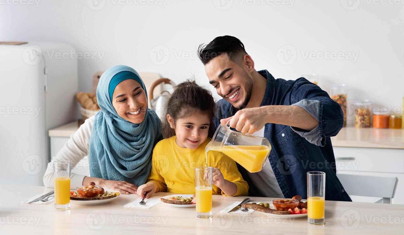 Portrait Of Happy Middle Eastern Family Of Three Having Breakfast In Kitchen Together, Arab Parents With Cute Little Daughter Enjoying Tasty Food And Drinking Orange Juice While Sitting At Table photo