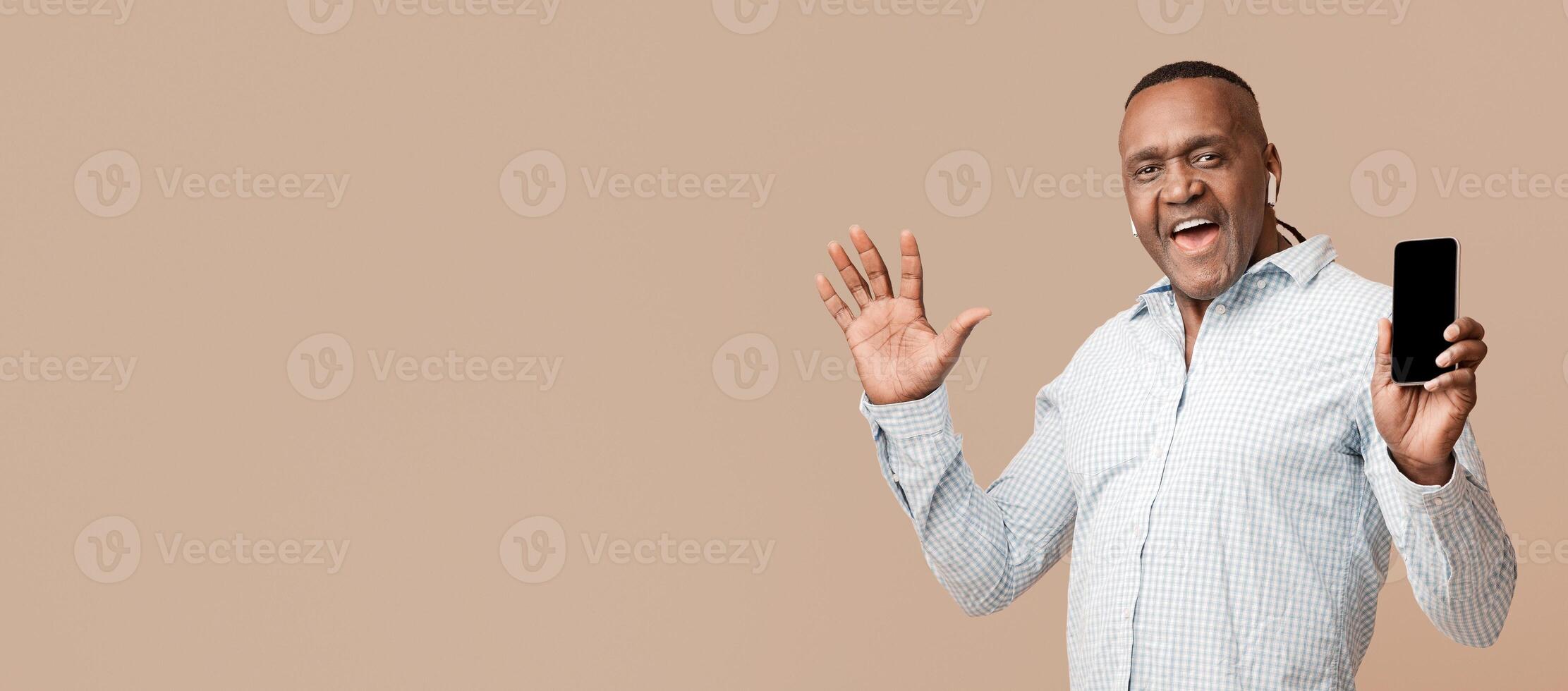 Music app. Positive mature african man in headphones enjoying music online, showing blank cellphone with mobile application photo