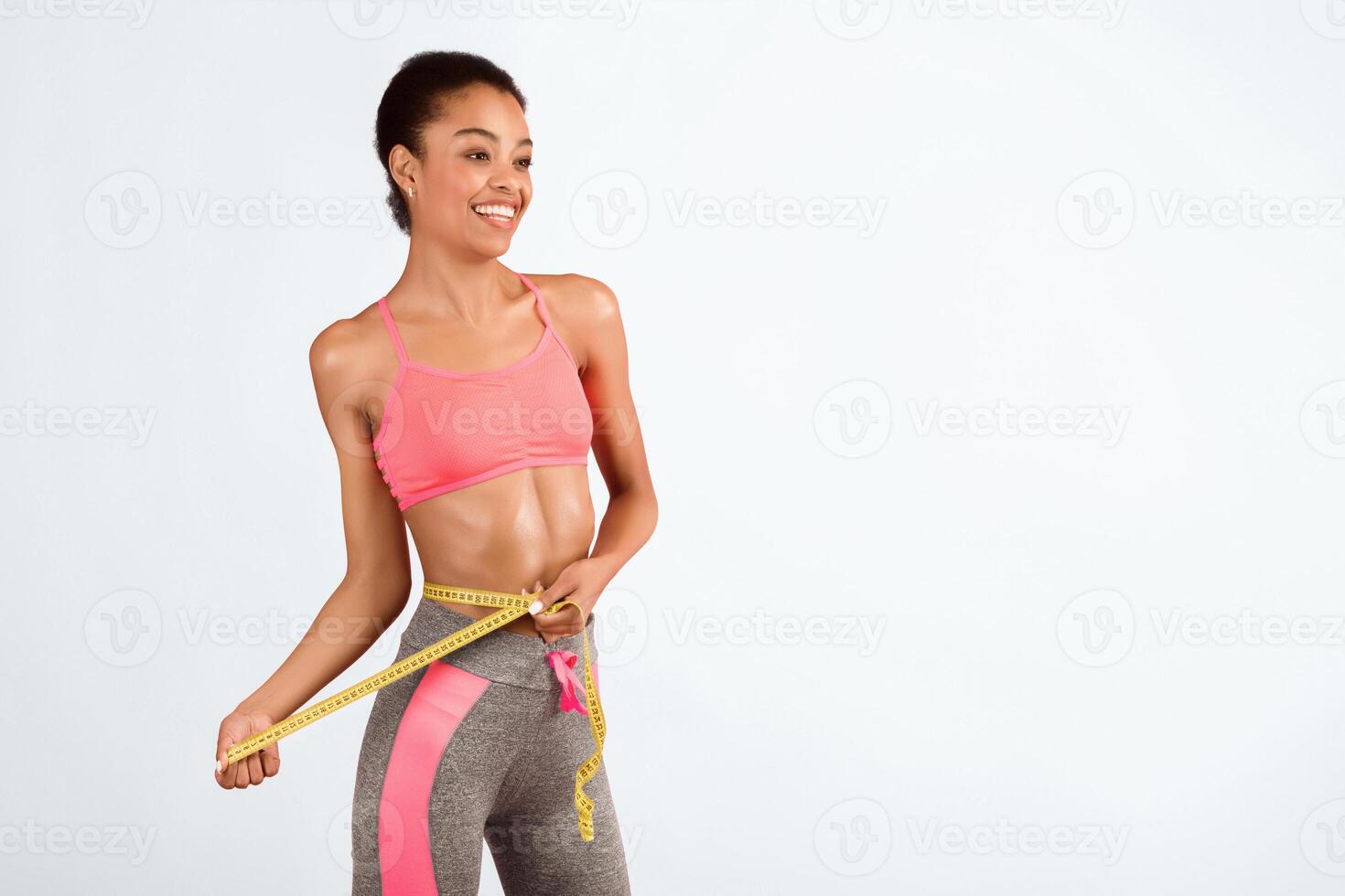 Losing Weight. Joyful Fit Afro Lady Measuring Waist With Tape Standing On Pink Studio Background. Empty Space photo