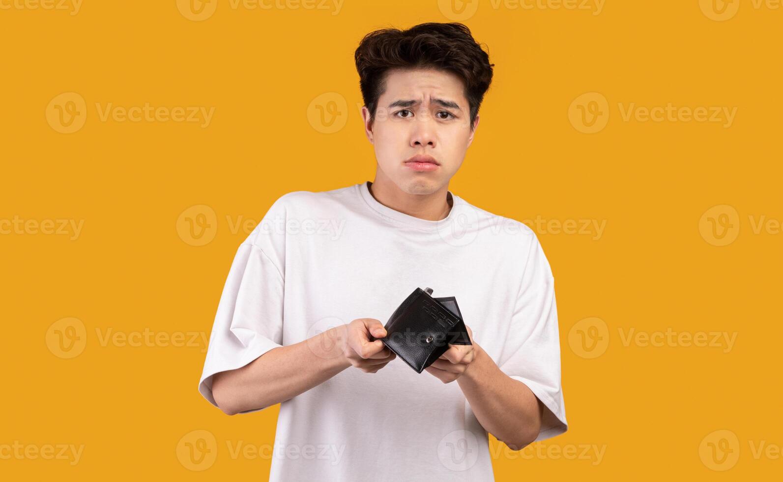I'm Broke. Portrait of sad asian bankrupt showing his empty wallet and looking at camera. Upset crying male model standing isolated over orange studio background wall. Poverty Concept. photo