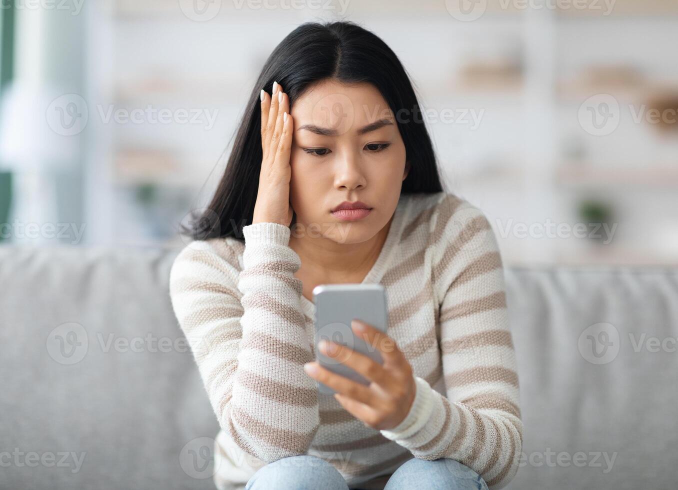 Bad News. Upset Asian Female Holding Smartphone, Looking At Mobile Phone Screen With Worry While Sitting On Couch At Home, Anxious Korean Woman Touching Head While Reading Unplesant Message, Closeup photo