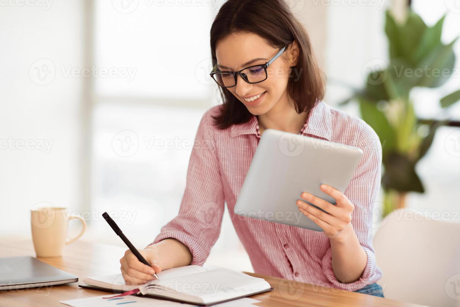 E-learning. Young woman in glasses holding tablet computer and writing with left hand in notebook, studying online photo