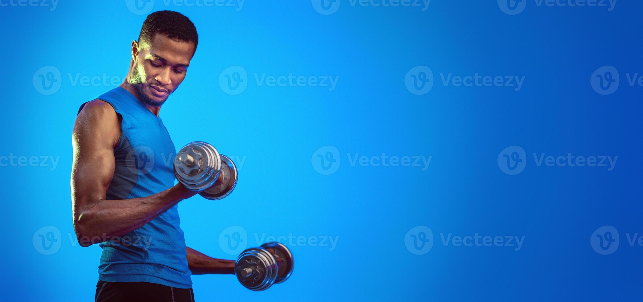 Strength Workout. Determined Fit Man Exercising With Dumbbells Pumping Bicep Muscles Over Blue Background. Studio Shot, Empty Space photo