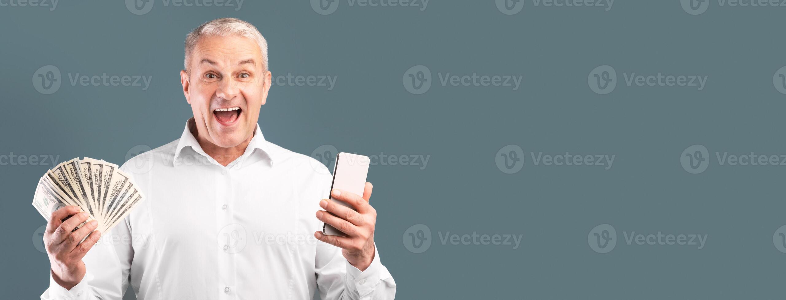 Making Money Via Internet. Excited mature man showing bunch of money, holding smartphone, free space, blue studio wall photo
