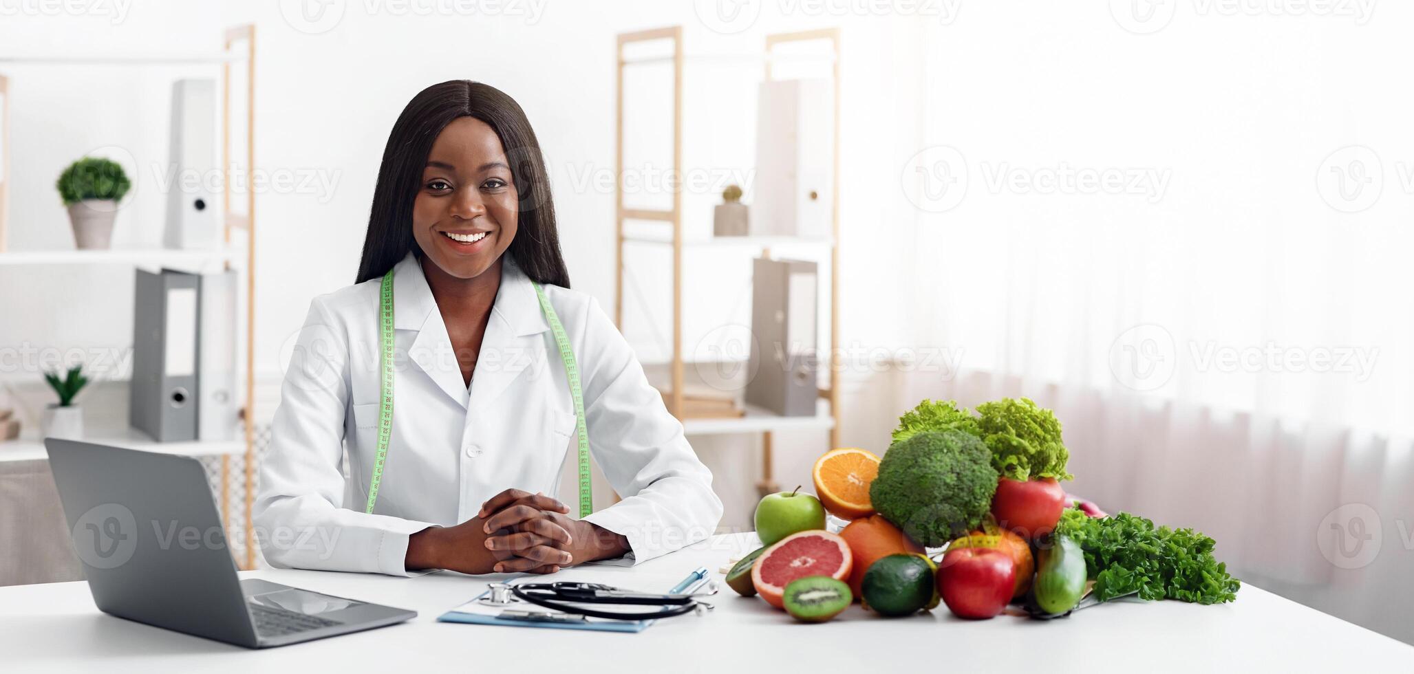 Smiling black lady doctor nutritionist sitting at workplace with vegetable and fruit, using laptop in office photo