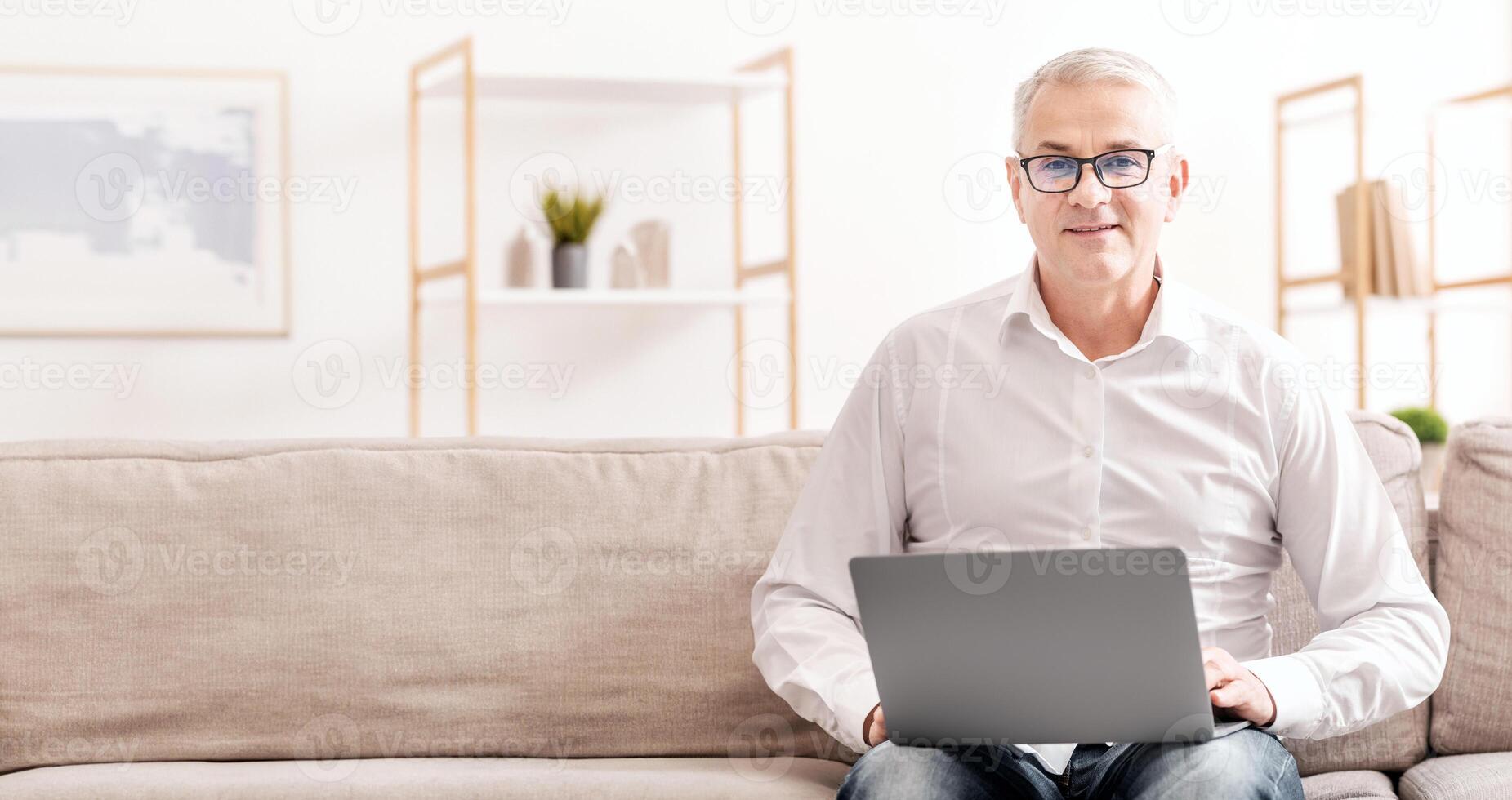 People And Technology Concept. Confident Mature Man Holding Computer On His Lap, Sitting On Couch, Free Space photo