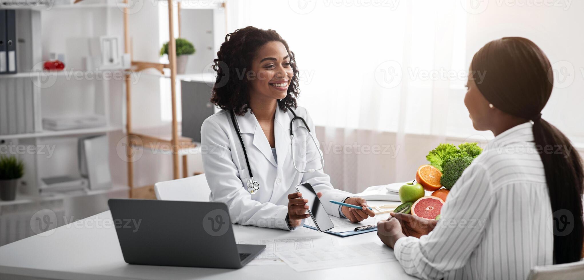 Cheerful dietologist showing female patient how to use mobile app for counting calories, clinic interior, free space photo