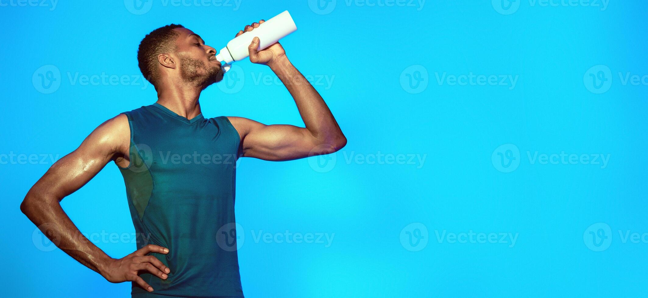 Workout Break. Black Sporty Guy Drinking Water After Exercising Over Blue Background In Studio. photo