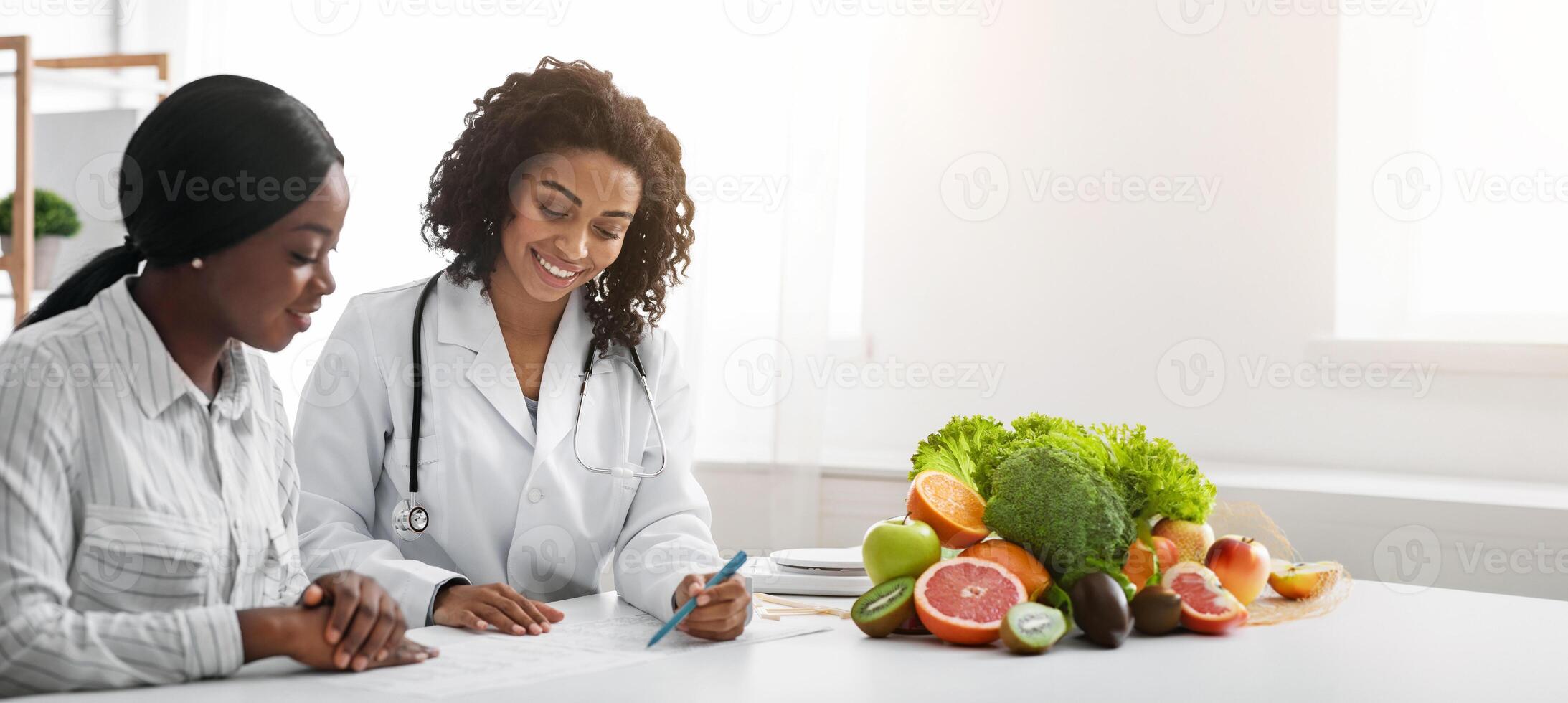 Cheerful african woman nutritionist making treatment plan for female patient, clinic interior photo