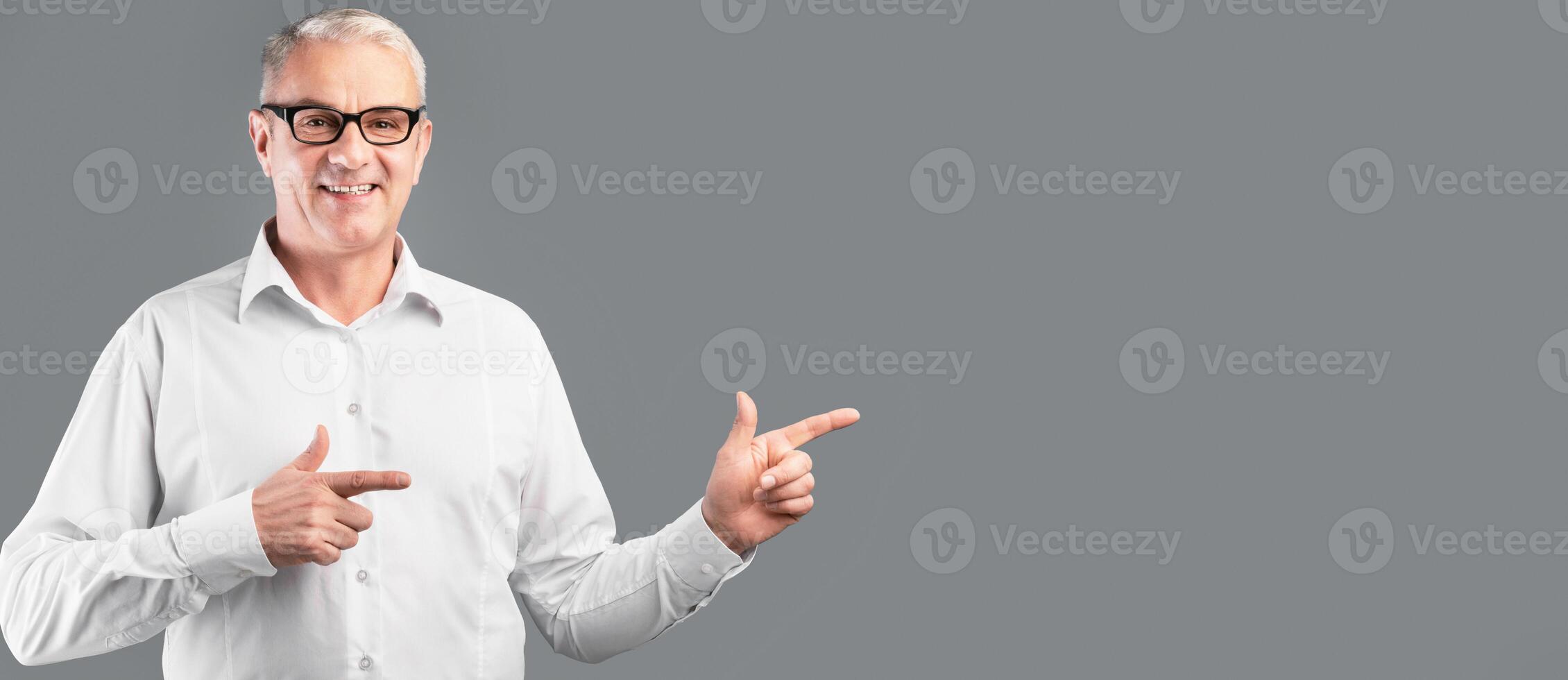 Cool Offer. Smiling mature man in glasses pointing two index fingers aside at copyspace, gray studio wall, panorama photo