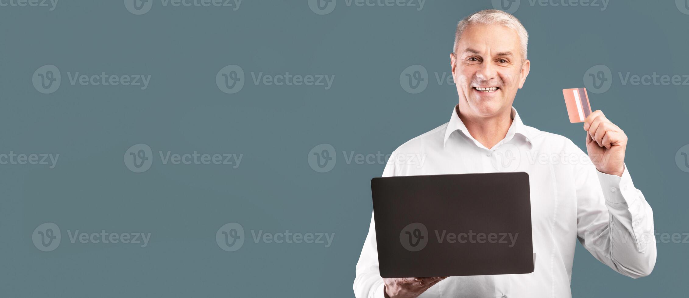 Easy Payment. Smiling middle-aged man showing credit card, holding laptop computer over blue studio wall photo