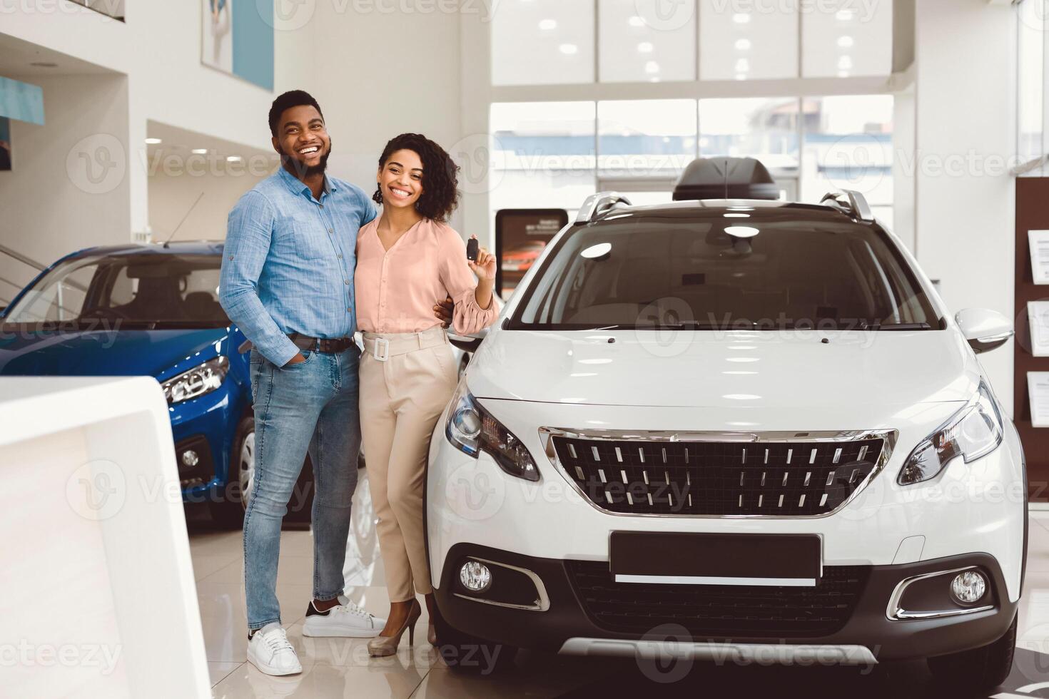 Car Buyers. Joyful Black Couple Hugging Showing Their New Auto Key Standing Near Automobile In Dealership Center. photo