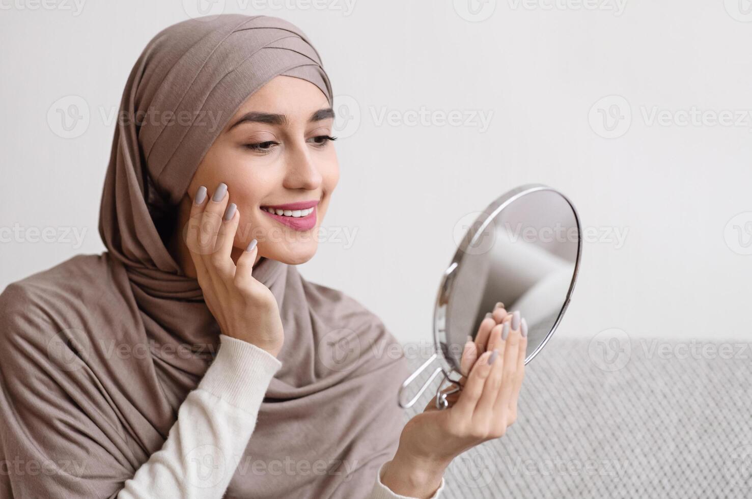 Beauty routine. Attractive muslim girl in hijab holding mirror and looking at her face with perfect soft skin, sitting on sofa at home, free space photo