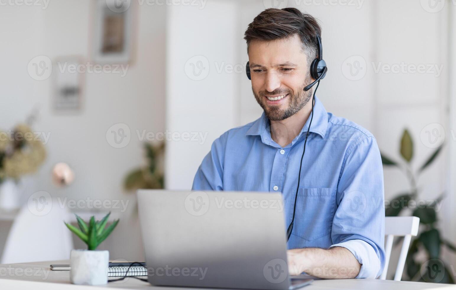 Call center operator at work. Positive male manager wearing headset and typing on laptop at workplace in modern office, consulting clients photo