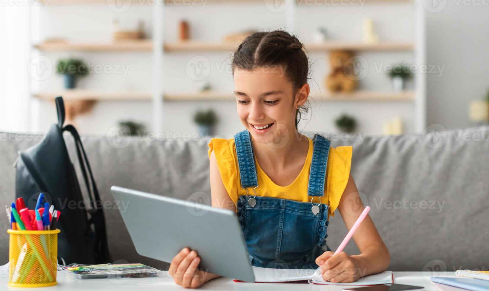 Children and Education Concept. Portrait of positive teen girl using doing homework writing in notebook sitting on couch at desk, holding and using tablet pad computer, enjoying distance homeschooling photo