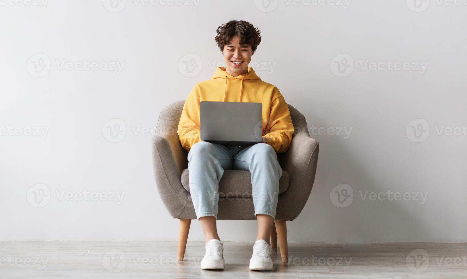 Happy young Asian male working online, sitting in armchair and using laptop against white studio wall, full length. Joyful millennial man surfing internet, watching webinar on portable computer photo